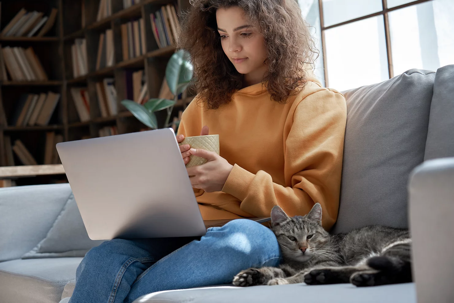 Assistante en ligne qui est en train de travailler de chez elle , elle a son ordinateur portable sur les genoux et un mug à la main, elle se trouve sur un canapé près d'une bibliothèque